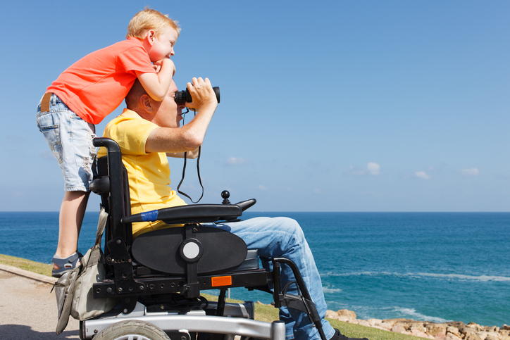 Father who utilizes a wheelchair looking through binoculars while his young boy stands behind him while hugging him looking in the distance. 