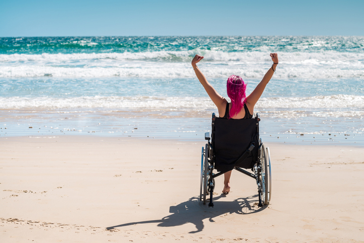 Young woman in the wheelchair at the beach enjoying fresh air with her arms wide open. 