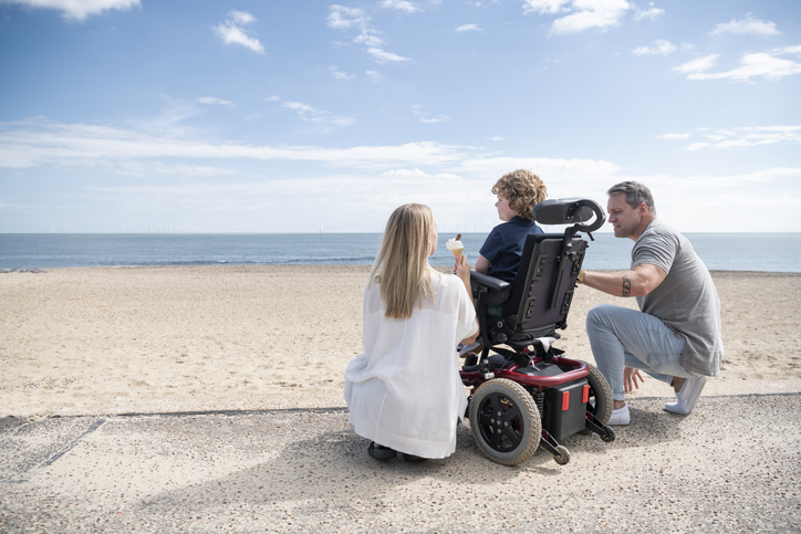 Mother and father kneeling by child, who is in wheelchair by the beach, enjoying an ice cream while looking at the waves.