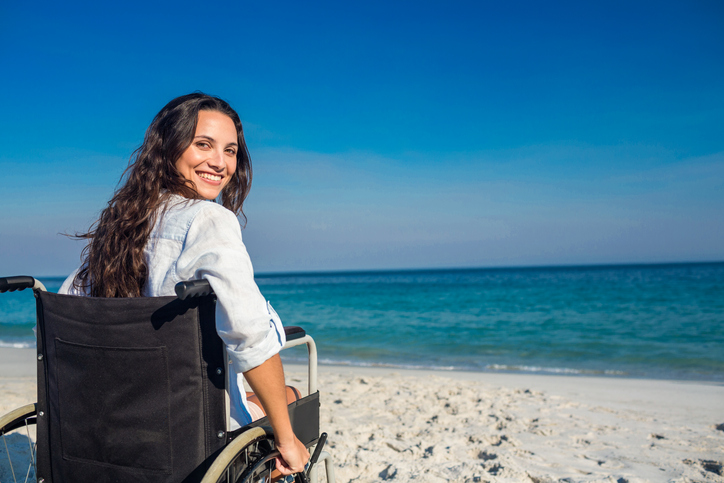 A young woman in wheelchair at the beach is looking back in to the camera and smiling 