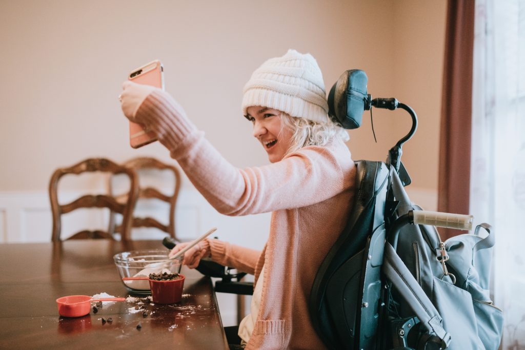 • A young woman in a wheelchair takes a selfie with her smartphone while mixing together ingredients for making chocolate chip cookies.